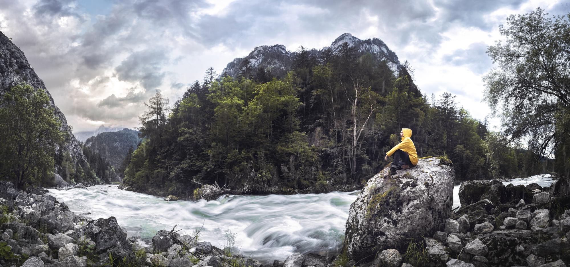 Ein Mann sitzt auf einem Felsen und sieht auf einen Fluss. Um ihn befinden sich Berge.