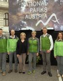 Eine Gruppe von acht Personen steht vor einer kleinen Bühe unter dem Slogan 'Nationalparks Austria'